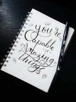 A spiral notebook with a motivational quote written in elegant, cursive handwriting. The phrase 'You're capable of amazing things' is prominently displayed. Accompanying the notebook is a black brush pen lying on the side. The notebook has a dotted background, adding to the artistic presentation.