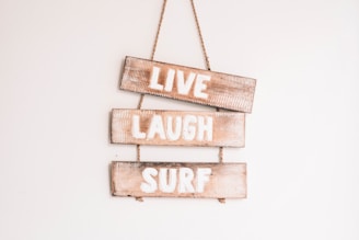 Three wooden signs are stacked vertically, each with a word painted in white. The words are 'LIVE', 'LAUGH', and 'SURF'. These signs are hanging on a wall and are connected by a rope.