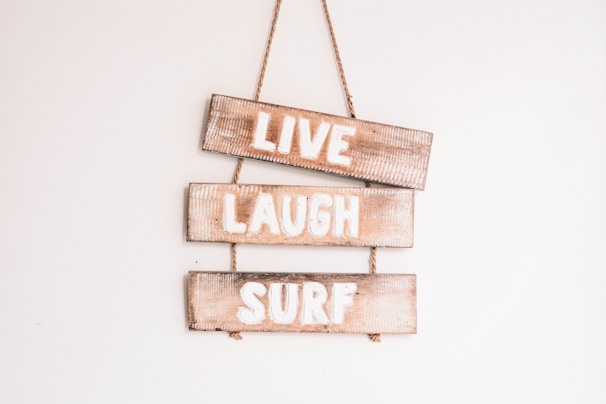 Three wooden signs are stacked vertically, each with a word painted in white. The words are 'LIVE', 'LAUGH', and 'SURF'. These signs are hanging on a wall and are connected by a rope.