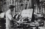 A person operates a large industrial machine in what appears to be a workshop or factory. The workspace is filled with complex metal machinery and various pieces of equipment. The individual is wearing a protective apron and focuses intently on the task, adjusting or controlling the machine. The environment is industrial and busy.