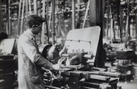 A person operates a large industrial machine in what appears to be a workshop or factory. The workspace is filled with complex metal machinery and various pieces of equipment. The individual is wearing a protective apron and focuses intently on the task, adjusting or controlling the machine. The environment is industrial and busy.
