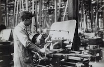 A person operates a large industrial machine in what appears to be a workshop or factory. The workspace is filled with complex metal machinery and various pieces of equipment. The individual is wearing a protective apron and focuses intently on the task, adjusting or controlling the machine. The environment is industrial and busy.