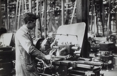 A person operates a large industrial machine in what appears to be a workshop or factory. The workspace is filled with complex metal machinery and various pieces of equipment. The individual is wearing a protective apron and focuses intently on the task, adjusting or controlling the machine. The environment is industrial and busy.