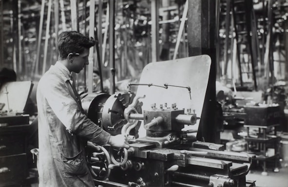 A person operates a large industrial machine in what appears to be a workshop or factory. The workspace is filled with complex metal machinery and various pieces of equipment. The individual is wearing a protective apron and focuses intently on the task, adjusting or controlling the machine. The environment is industrial and busy.
