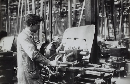 A person operates a large industrial machine in what appears to be a workshop or factory. The workspace is filled with complex metal machinery and various pieces of equipment. The individual is wearing a protective apron and focuses intently on the task, adjusting or controlling the machine. The environment is industrial and busy.