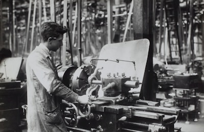 A person operates a large industrial machine in what appears to be a workshop or factory. The workspace is filled with complex metal machinery and various pieces of equipment. The individual is wearing a protective apron and focuses intently on the task, adjusting or controlling the machine. The environment is industrial and busy.