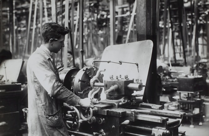 A person operates a large industrial machine in what appears to be a workshop or factory. The workspace is filled with complex metal machinery and various pieces of equipment. The individual is wearing a protective apron and focuses intently on the task, adjusting or controlling the machine. The environment is industrial and busy.