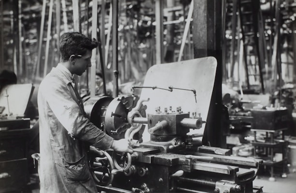 A person operates a large industrial machine in what appears to be a workshop or factory. The workspace is filled with complex metal machinery and various pieces of equipment. The individual is wearing a protective apron and focuses intently on the task, adjusting or controlling the machine. The environment is industrial and busy.