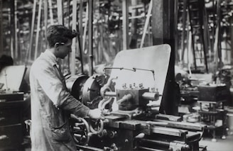 A person operates a large industrial machine in what appears to be a workshop or factory. The workspace is filled with complex metal machinery and various pieces of equipment. The individual is wearing a protective apron and focuses intently on the task, adjusting or controlling the machine. The environment is industrial and busy.