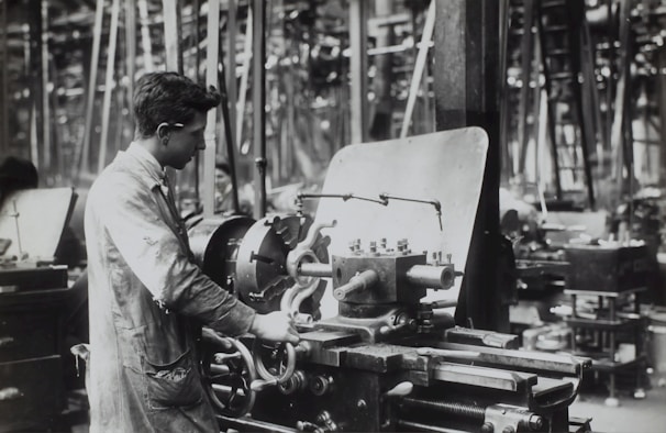 A person operates a large industrial machine in what appears to be a workshop or factory. The workspace is filled with complex metal machinery and various pieces of equipment. The individual is wearing a protective apron and focuses intently on the task, adjusting or controlling the machine. The environment is industrial and busy.