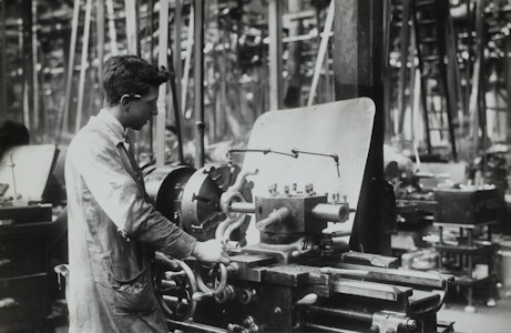 A person operates a large industrial machine in what appears to be a workshop or factory. The workspace is filled with complex metal machinery and various pieces of equipment. The individual is wearing a protective apron and focuses intently on the task, adjusting or controlling the machine. The environment is industrial and busy.