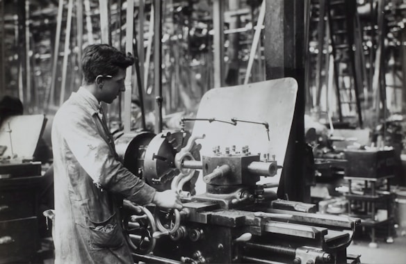 A person operates a large industrial machine in what appears to be a workshop or factory. The workspace is filled with complex metal machinery and various pieces of equipment. The individual is wearing a protective apron and focuses intently on the task, adjusting or controlling the machine. The environment is industrial and busy.