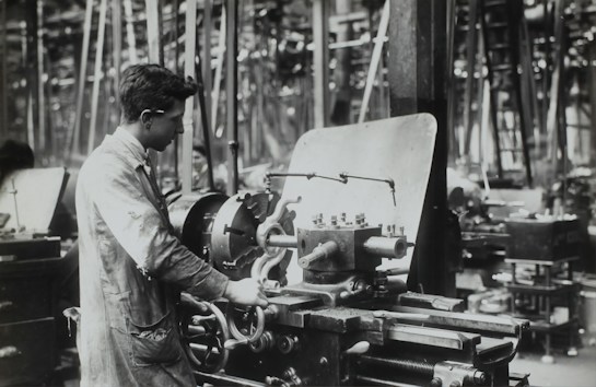 A person operates a large industrial machine in what appears to be a workshop or factory. The workspace is filled with complex metal machinery and various pieces of equipment. The individual is wearing a protective apron and focuses intently on the task, adjusting or controlling the machine. The environment is industrial and busy.