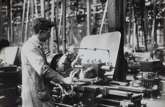A person operates a large industrial machine in what appears to be a workshop or factory. The workspace is filled with complex metal machinery and various pieces of equipment. The individual is wearing a protective apron and focuses intently on the task, adjusting or controlling the machine. The environment is industrial and busy.