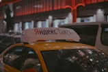 A Yandex Taxi is parked in a dimly lit urban area at night. The car is yellow with a white top sign featuring Cyrillic text. Red and white lights from nearby buildings create a warm glow in the background. Several vehicles are parked around the taxi, indicating a busy street setting.