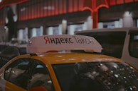 A Yandex Taxi is parked in a dimly lit urban area at night. The car is yellow with a white top sign featuring Cyrillic text. Red and white lights from nearby buildings create a warm glow in the background. Several vehicles are parked around the taxi, indicating a busy street setting.