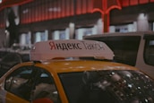 A Yandex Taxi is parked in a dimly lit urban area at night. The car is yellow with a white top sign featuring Cyrillic text. Red and white lights from nearby buildings create a warm glow in the background. Several vehicles are parked around the taxi, indicating a busy street setting.