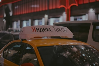 A Yandex Taxi is parked in a dimly lit urban area at night. The car is yellow with a white top sign featuring Cyrillic text. Red and white lights from nearby buildings create a warm glow in the background. Several vehicles are parked around the taxi, indicating a busy street setting.
