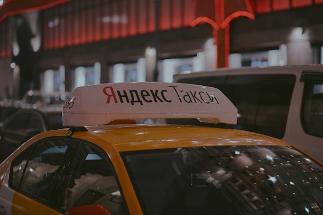 A Yandex Taxi is parked in a dimly lit urban area at night. The car is yellow with a white top sign featuring Cyrillic text. Red and white lights from nearby buildings create a warm glow in the background. Several vehicles are parked around the taxi, indicating a busy street setting.