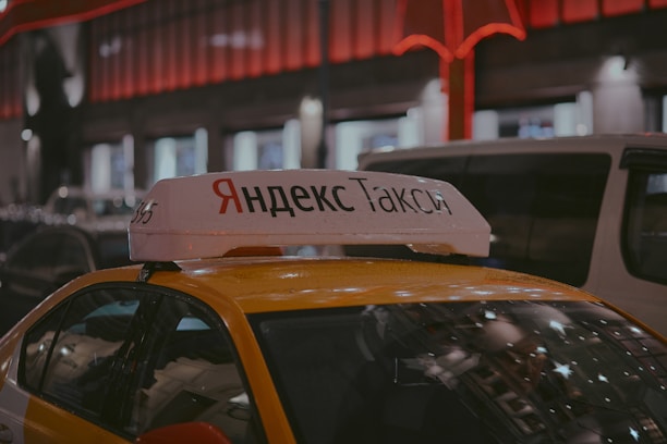 A Yandex Taxi is parked in a dimly lit urban area at night. The car is yellow with a white top sign featuring Cyrillic text. Red and white lights from nearby buildings create a warm glow in the background. Several vehicles are parked around the taxi, indicating a busy street setting.