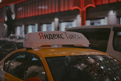 A Yandex Taxi is parked in a dimly lit urban area at night. The car is yellow with a white top sign featuring Cyrillic text. Red and white lights from nearby buildings create a warm glow in the background. Several vehicles are parked around the taxi, indicating a busy street setting.