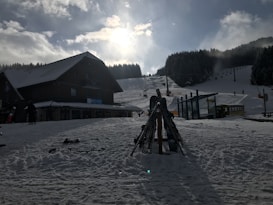A snowy landscape with a rustic wooden building on the left. In front of the building, stacks of skis are grouped together. The background features a ski slope with several people skiing and a ski lift ascending the hill. Tall evergreen trees border the snowy hill, and the sun shines through partly cloudy skies.