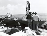 Several individuals are gathered around equipment on a vessel at sea. The equipment includes metallic structures and what appears to be a surveying device. A sign reads 'KEEP OFF.' The ocean and a slightly cloudy sky are visible in the background.