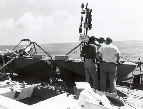 Several individuals are gathered around equipment on a vessel at sea. The equipment includes metallic structures and what appears to be a surveying device. A sign reads 'KEEP OFF.' The ocean and a slightly cloudy sky are visible in the background.