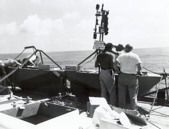 Several individuals are gathered around equipment on a vessel at sea. The equipment includes metallic structures and what appears to be a surveying device. A sign reads 'KEEP OFF.' The ocean and a slightly cloudy sky are visible in the background.