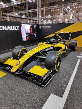 A bright yellow Renault Formula 1 car is displayed in a well-lit exhibition space, with branding and logos clearly visible on the vehicle's body. The background features a large screen with a person displayed on it, alongside more Renault Sport branding. The environment is modern, with industrial lighting fixtures and a structured metal framework.