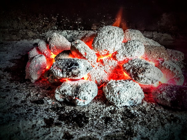 A collection of glowing charcoal briquettes surrounded by ash, emitting a soft, warm glow. They are arranged in a pile with bright orange and red embers visible between them, suggesting intense heat.
