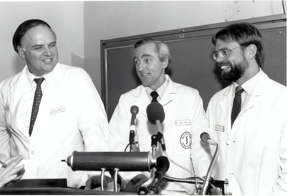 três homens sorridentes vestindo vestes em frente aos microfones