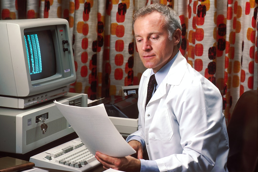 man reading papers in front of computer, Doctor sitting at his office desk accessing PDQ on his computer in the 1980s. The Physicians Data Query was designed by the National Cancer Institute to help physicians obtain information about the most up-to-date protocols, physicians, and clinics treating cancer patients.<p></figure>1987