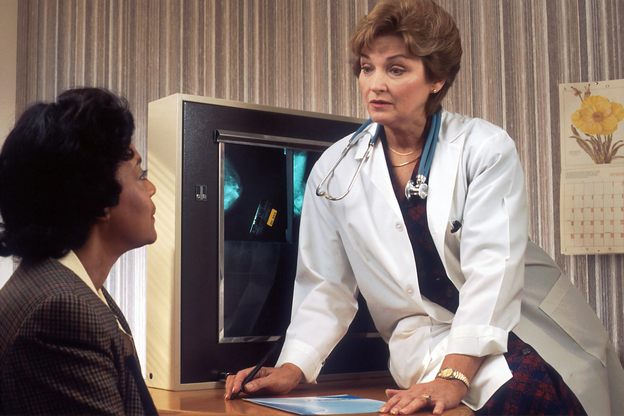 A Caucasian female doctor, sitting on her desk with mammograms on a view box behind her, speaks with an African-American female patient. 1990