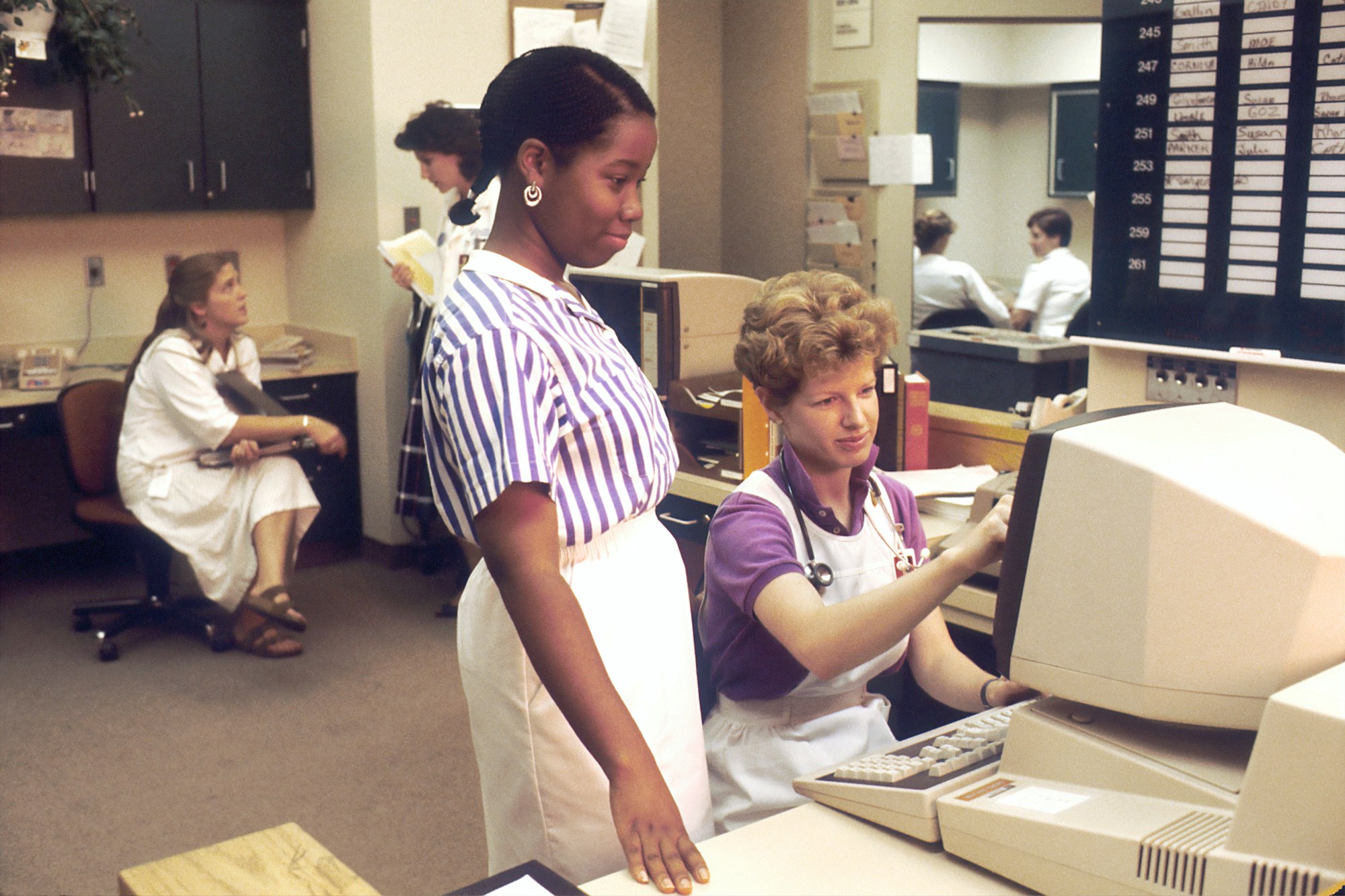 	Several nurses in a nurses' station, two of which are working on a computer accessing PDQ. Physicians Data Query was designed by the National Cancer Institute to help physicians and patients obtain information on protocols, physician referrals and clinics using the most up-to-date cancer treatments. 1987