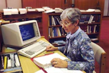 An elderly woman typing thoughtfully on a laptop in a sunlit room filled with books.