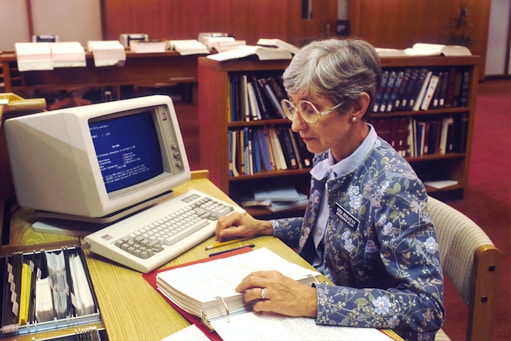 An older woman is sitting at a desk typing on a vintage computer keyboard with a monochrome monitor. She wears glasses and a floral-patterned jacket. A binder with papers is open in front of her, and several open books are in the background, suggesting a library or office environment.