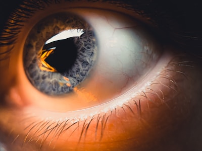 A close-up view of a human eye showcasing intricate details of the iris and pupil. The eye is illuminated with a warm light that highlights the organic textures and colors surrounding the iris. Fine eyelashes are visible along the edge of the eyelid.