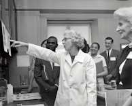 A group of people in a professional setting with a woman in a lab coat at the forefront, pointing towards some papers. The individuals are engaged and appear to be listening intently. The room resembles a laboratory or office space, as suggested by the calendar on the wall and some lab equipment on the table.