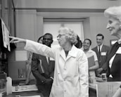 A group of people in a professional setting with a woman in a lab coat at the forefront, pointing towards some papers. The individuals are engaged and appear to be listening intently. The room resembles a laboratory or office space, as suggested by the calendar on the wall and some lab equipment on the table.