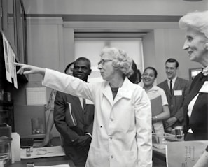 A group of people in a professional setting with a woman in a lab coat at the forefront, pointing towards some papers. The individuals are engaged and appear to be listening intently. The room resembles a laboratory or office space, as suggested by the calendar on the wall and some lab equipment on the table.