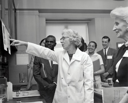 A group of people in a professional setting with a woman in a lab coat at the forefront, pointing towards some papers. The individuals are engaged and appear to be listening intently. The room resembles a laboratory or office space, as suggested by the calendar on the wall and some lab equipment on the table.