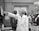 A group of people in a professional setting with a woman in a lab coat at the forefront, pointing towards some papers. The individuals are engaged and appear to be listening intently. The room resembles a laboratory or office space, as suggested by the calendar on the wall and some lab equipment on the table.