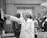 A group of people in a professional setting with a woman in a lab coat at the forefront, pointing towards some papers. The individuals are engaged and appear to be listening intently. The room resembles a laboratory or office space, as suggested by the calendar on the wall and some lab equipment on the table.