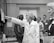 A group of people in a professional setting with a woman in a lab coat at the forefront, pointing towards some papers. The individuals are engaged and appear to be listening intently. The room resembles a laboratory or office space, as suggested by the calendar on the wall and some lab equipment on the table.