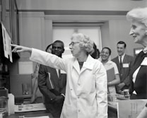A group of people in a professional setting with a woman in a lab coat at the forefront, pointing towards some papers. The individuals are engaged and appear to be listening intently. The room resembles a laboratory or office space, as suggested by the calendar on the wall and some lab equipment on the table.