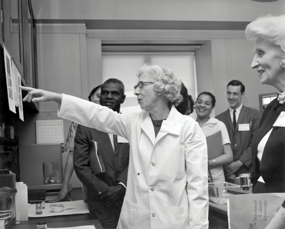 A group of people in a professional setting with a woman in a lab coat at the forefront, pointing towards some papers. The individuals are engaged and appear to be listening intently. The room resembles a laboratory or office space, as suggested by the calendar on the wall and some lab equipment on the table.