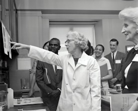 A group of people in a professional setting with a woman in a lab coat at the forefront, pointing towards some papers. The individuals are engaged and appear to be listening intently. The room resembles a laboratory or office space, as suggested by the calendar on the wall and some lab equipment on the table.