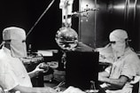 Two individuals dressed in protective clothing and masks are working in a laboratory setting. They are seated on either side of a large distillation apparatus, which includes various glassware such as a round-bottom flask. The environment suggests a high level of precision and attention to cleanliness and safety.