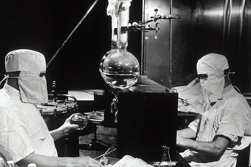 Two individuals dressed in protective clothing and masks are working in a laboratory setting. They are seated on either side of a large distillation apparatus, which includes various glassware such as a round-bottom flask. The environment suggests a high level of precision and attention to cleanliness and safety.